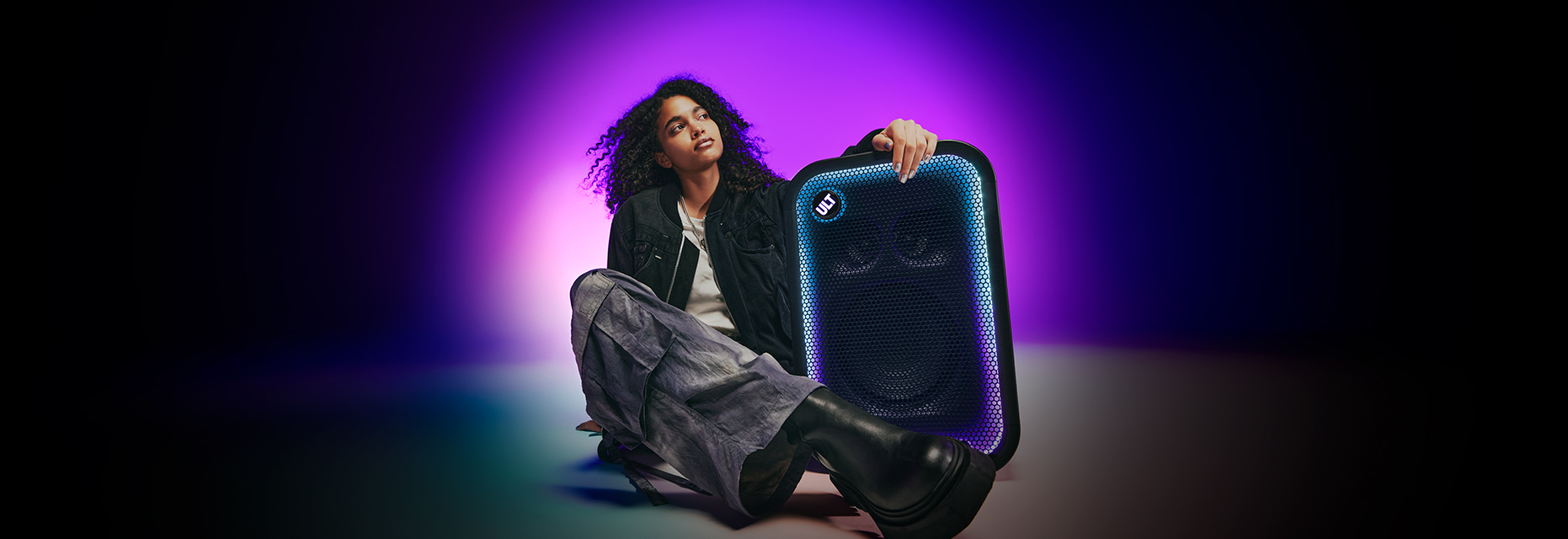 Photograph of a person with long curly dark hair sitting on the floor against a purple gradient background. Wearing a black jacket and grey trousers, the person rests a hand on a ULT TOWER 5 speaker bordered with glowing blue and purple LED lights and a circular ULT logo.